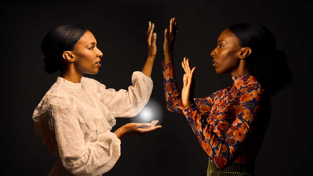 two figures (same person dressed to represent two different people). They are facing into eachother. One set of their palms are mirroring each other. The ancestral fiigure (in white on the left) is holding a white light in her other palm